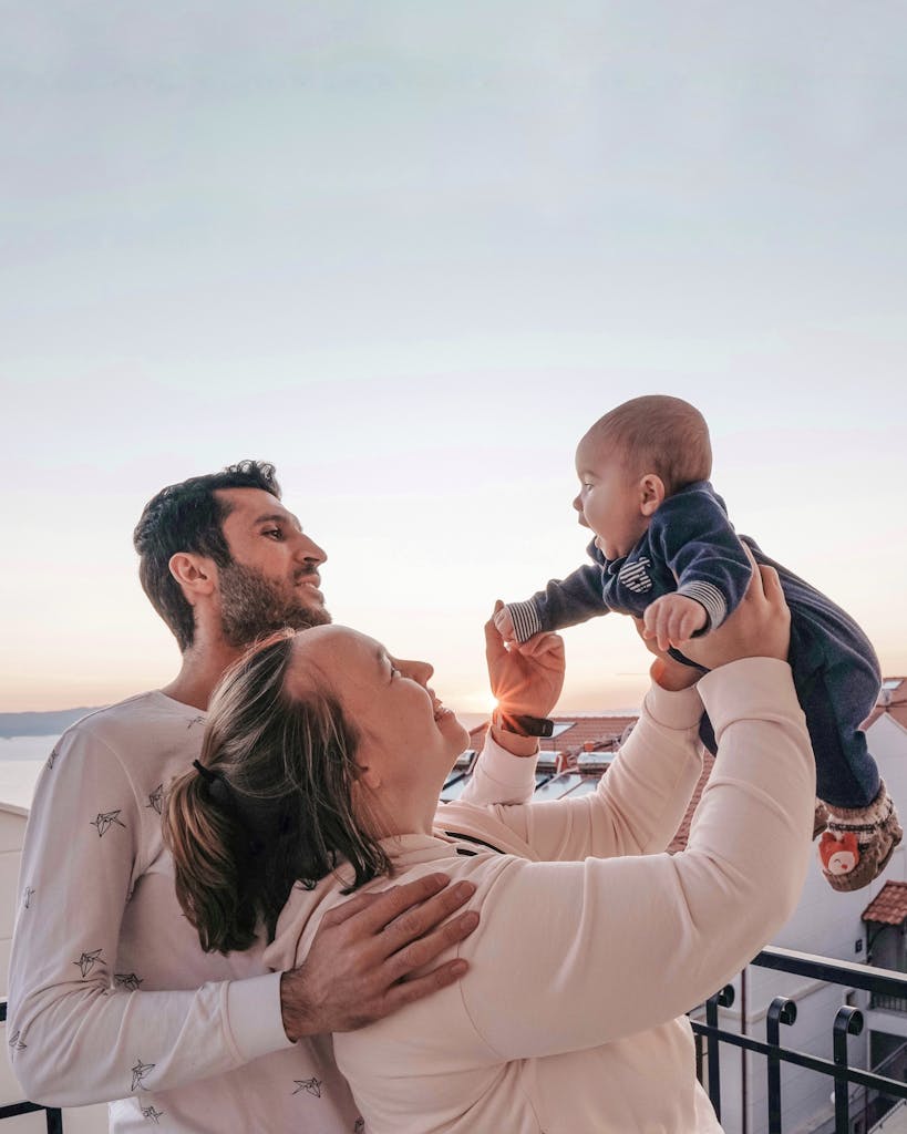 Side view of happy grandma raising adorable baby while standing near bearded ethnic son on fenced terrace in apartment