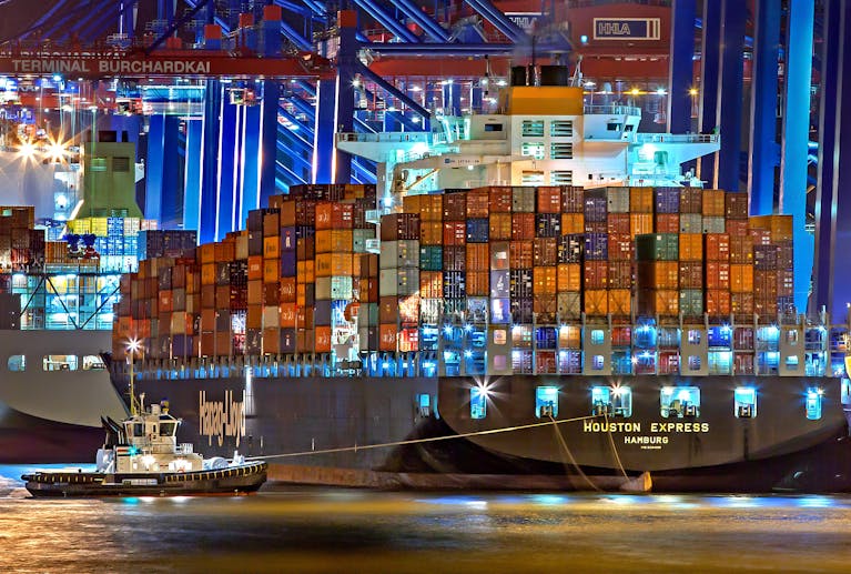 A brightly lit cargo ship at Hamburg harbor with stacked containers and a tugboat.