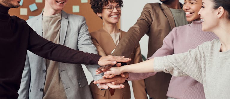 A group of happy, diverse colleagues celebrating teamwork and cooperation with a group high five indoors.