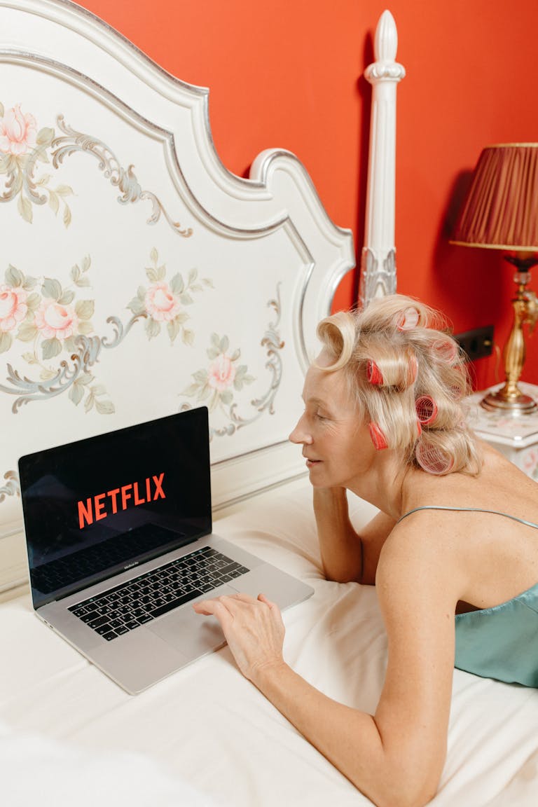 Senior woman with curlers enjoying Netflix on a laptop while lying in a stylish bedroom.